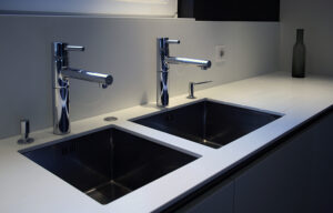 Salle de bain sur mesure réalisée par LDC Agencement – L’Arche du Bois : deux lavabos noirs avec robinets modernes, plan de travail et mur blancs, bouteille posée en haut à droite. Vasque sur mesure Alsace