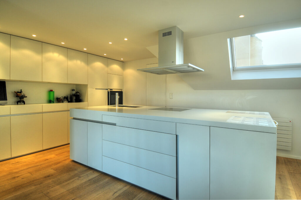 Cuisine sur mesure par LDC Agencement – L’Arche du Bois : îlot central blanc avec lavabo et plaque de cuisson intégrés, tiroirs intégrés, placards et étagères à gauche et fenêtre avec hotte au fond.
