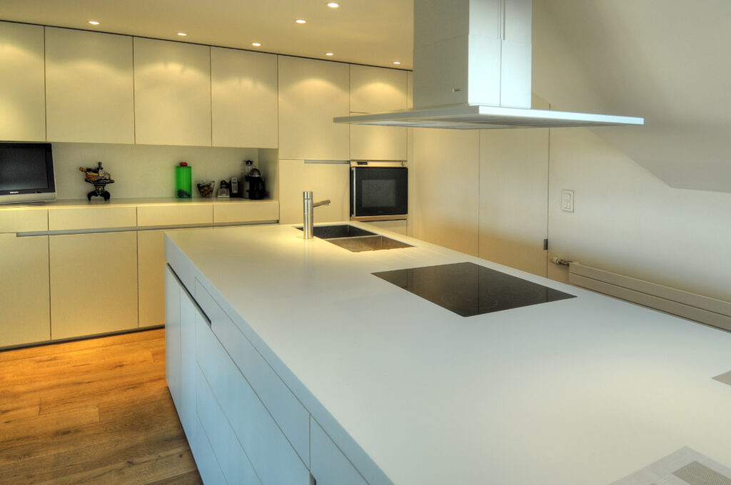 Cuisine sur mesure par LDC Agencement – L’Arche du Bois : îlot central blanc avec lavabo et plaque de cuisson intégrés, tiroirs intégrés, placards et étagères à gauche et fenêtre avec hotte au fond.