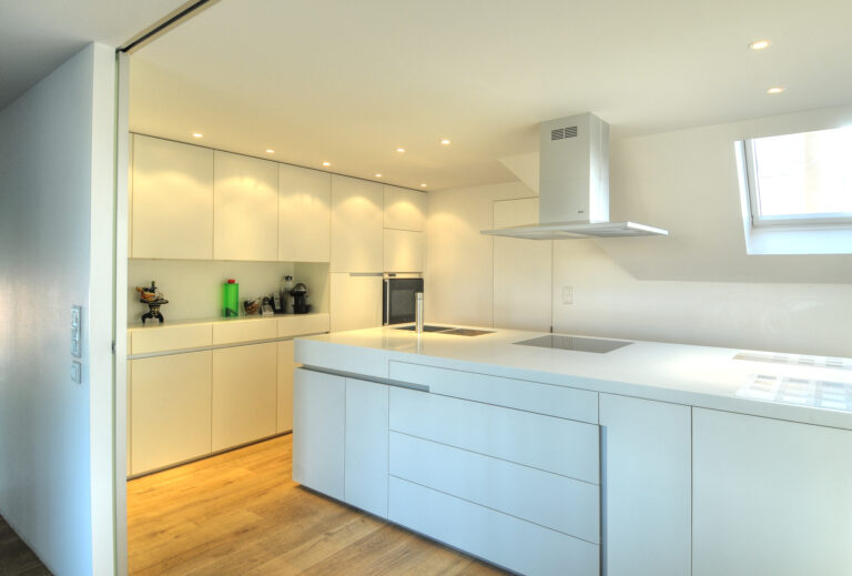 Cuisine sur mesure par LDC Agencement – L’Arche du Bois : îlot central blanc avec lavabo et plaque de cuisson intégrés, tiroirs intégrés, placards et étagères à gauche et fenêtre avec hotte au fond.