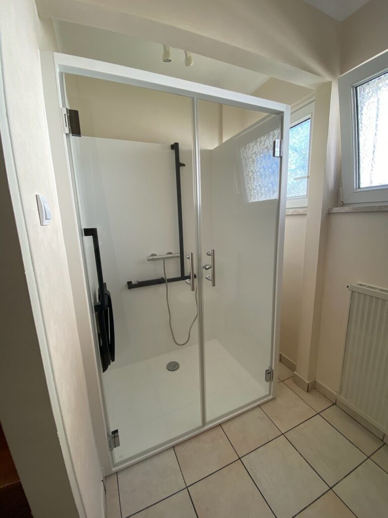 Cabine de douche Danaé by L'Arche du Bois avec double porte vitrée fermée, laissant voir le siège PMR et les barres de maintien à l'intérieur.