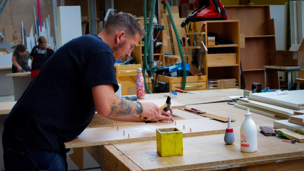 Ouvrier de L’Arche du Bois utilisant un marteau pour fixer des chevilles en bois sur une planche, avec plusieurs planches devant lui dans l’atelier de fabrication.
