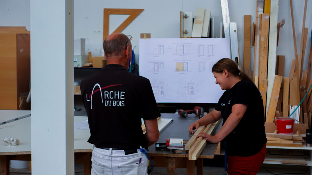 Deux techniciens de L’Arche du Bois discutant d’un projet dans l’atelier, avec un plan affiché derrière eux. L’un porte un t-shirt avec le logo de L’Arche du Bois.