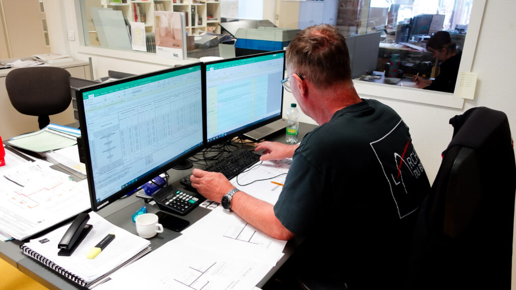 Ingénieur du bureau d’étude de L’Arche du Bois travaillant sur des plans de projet, devant deux écrans affichant des tableaux Excel, portant un t-shirt avec le logo de L’Arche du Bois, entouré de documents techniques et plans d’architecture en bois.