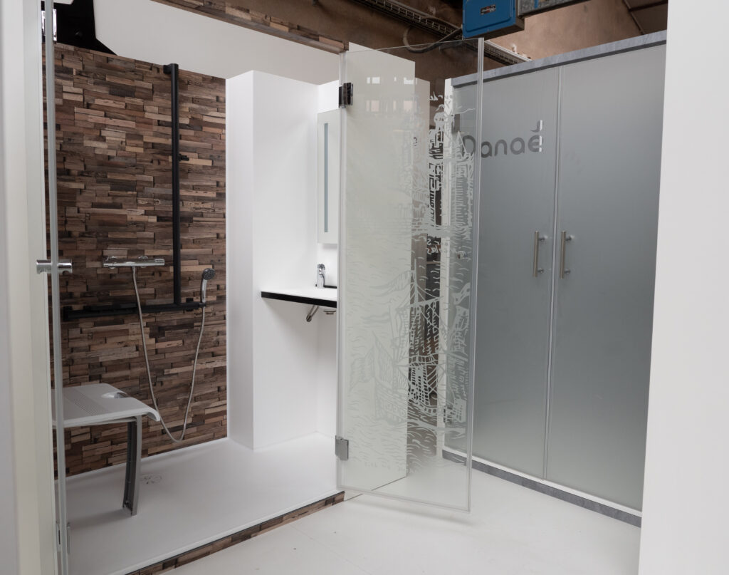 Showroom de la douche accessible Danaé by L'Arche du Bois, avec porte ouverte montrant un siège PMR, une barre de maintien et un panneau mural finition bois rustique.