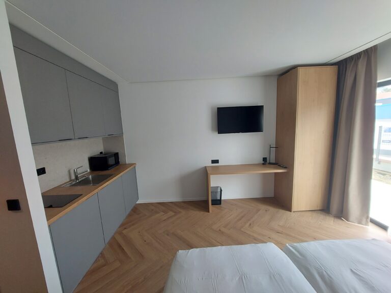 agencement hôtelier sur-mesure, Vue d'ensemble d'un studio hôtelier moderne avec une kitchenette grise à gauche et un ensemble bureau-penderie en bois clair à droite sur un parquet en chevron.