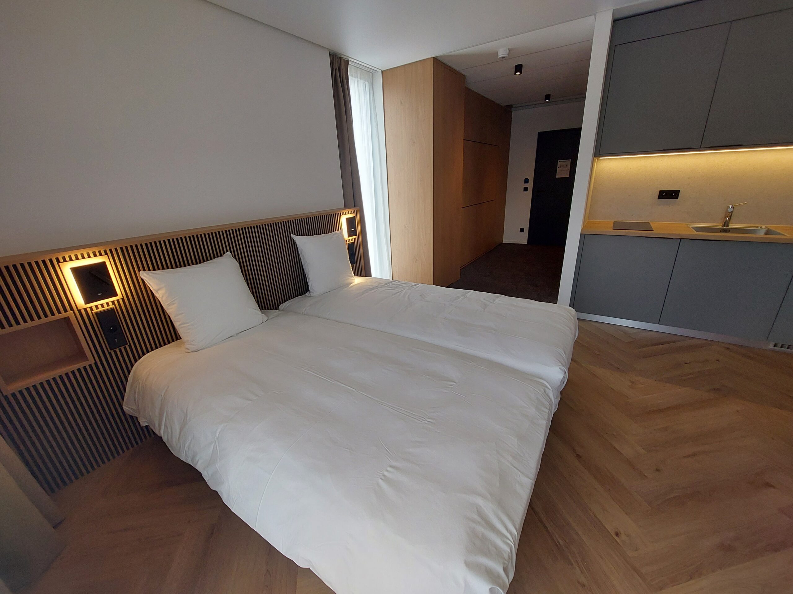 agencement hôtelier sur-mesure, Perspective depuis le pied du lit montrant la kitchenette grise intégrée et le bureau en bois clair, dans une chambre lumineuse.