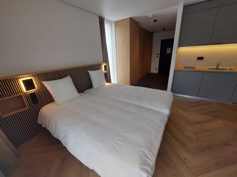 Chambre d'hôtel avec un lit double à tête de lit en tasseaux de bois, une kitchenette moderne grise et un sol en parquet en chevrons.