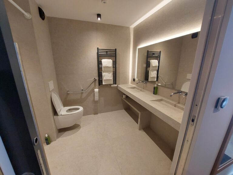 Grande salle de bain avec un double plan vasque suspendu en pierre beige, un large miroir rétroéclairé et des équipements PMR incluant des barres d'appui chromées.