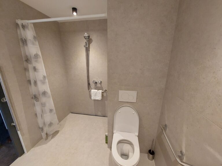 Salle de bain moderne et accessible avec carrelage beige effet pierre, douche à l'italienne sans seuil avec barre de maintien et WC suspendu.