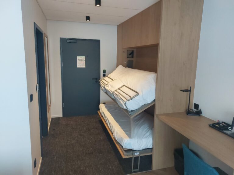 Vue d'un système de lits superposés escamotables en cours d'ouverture, intégré dans un grand meuble en bois clair avec un bureau attenant.