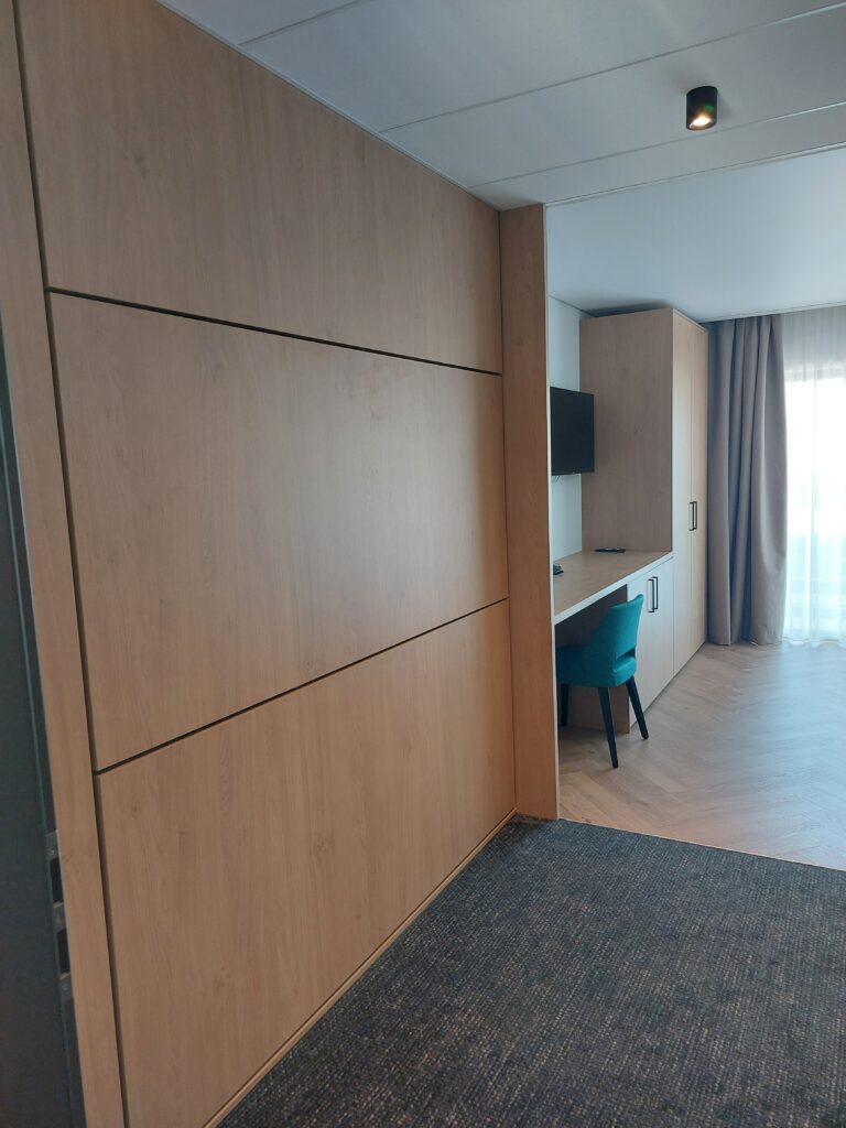 Aménagement sur mesure en bois clair avec lit escamotable refermé formant une paroi murale lisse, à côté d'un espace bureau et de rangements coordonnés.