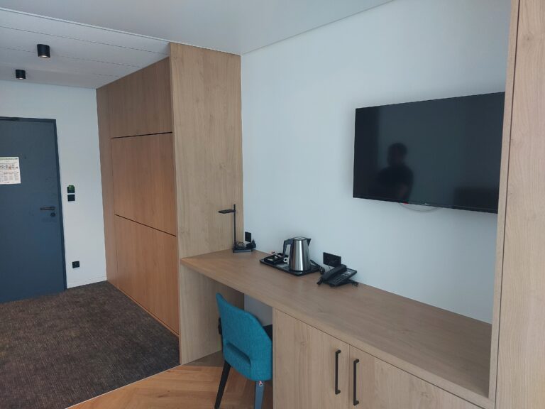 agencement hôtelier sur-mesure, Plan de travail en bois clair prolongé par un meuble bas, avec une chaise bleue, devant un mur blanc équipé d'une télévision et d'un lit escamotable fermé en arrière-plan.