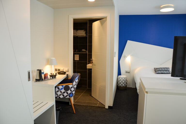 Intérieur d'une chambre d'hôtel moderne avec un bureau blanc, une tête de lit géométrique "origami" sur un mur bleu, et un meuble TV minimaliste.