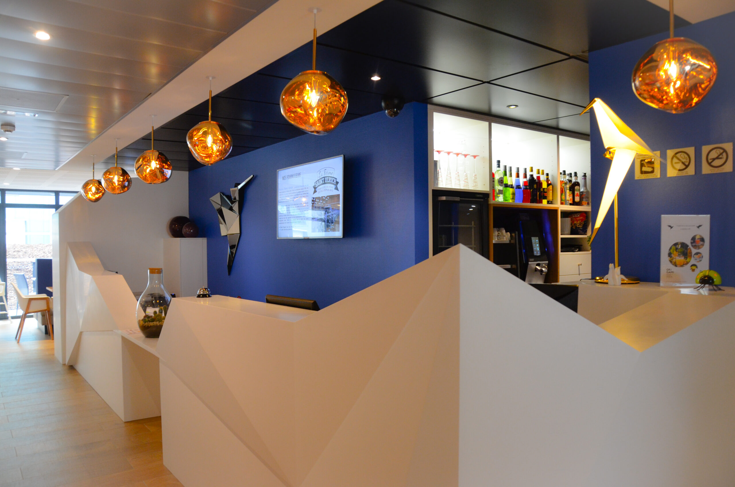 Vue large de la réception d'un hôtel avec un imposant comptoir blanc facetté, des luminaires suspendus et un bar en arrière-plan.