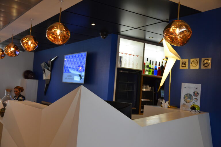 Banque d'accueil blanche aux formes géométriques facettées (style origami) devant un mur bleu profond avec des suspensions lumineuses cuivrées.