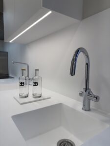 Détail d'un évier blanc épuré avec robinetterie chromée et bouteilles siglées EB dans une cuisine professionnelle au design minimaliste pour le projet cuisine Bugatti.