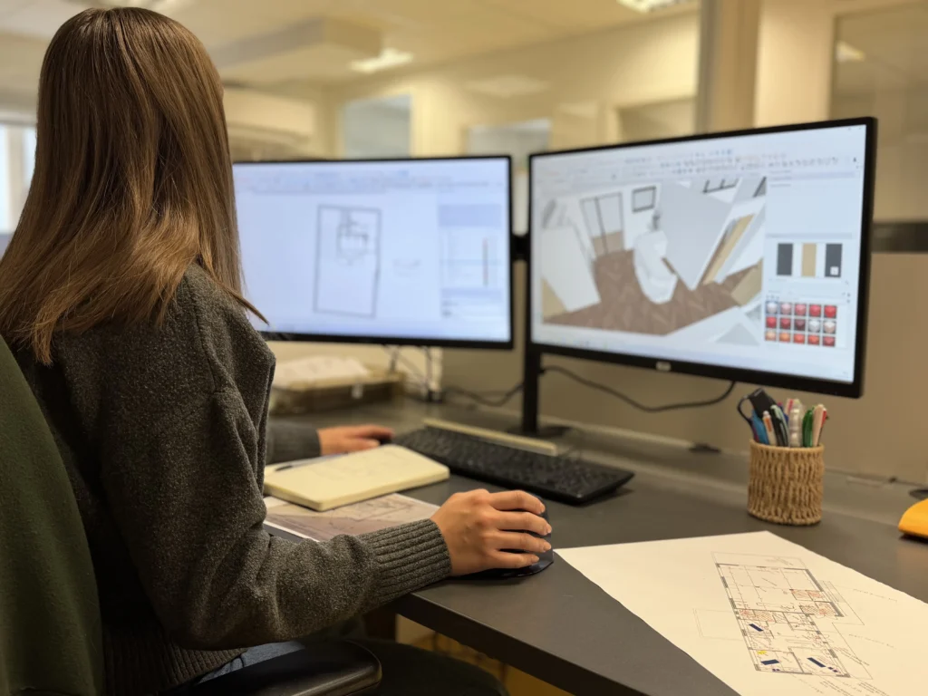 Vue de dos d'une conceptrice travaillant à un bureau d'angle moderne avec deux grands moniteurs. L'un montre des plans techniques CAO, l'autre un rendu 3D d'un aménagement intérieur. Clavier, souris, plans papier et pot à crayons sur la table. Menuiserie et Agencement Intérieur