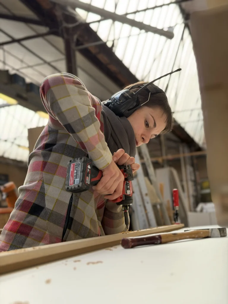 Gros plan sur une jeune menuisière portant des protections auditives, concentrée sur son travail. Elle utilise une visseuse/perceuse sans fil pour visser un élément en bois pré-usiné. Veste à carreaux. Scène d'atelier d'agencement.