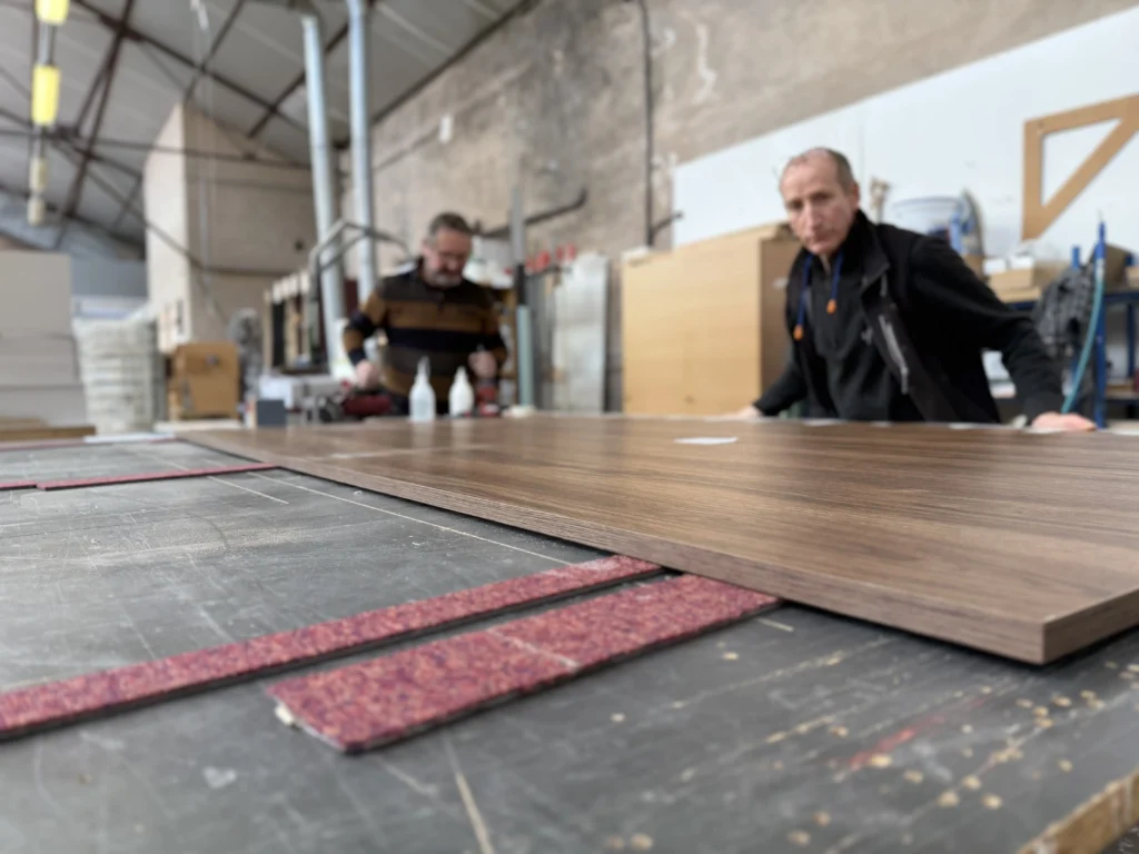 Deux artisans menuisiers travaillent dans un grand atelier industriel lumineux. Un grand panneau de bois plaqué est maintenu sur un établi avec des patins rouges et des guides de précision. Outils, machines et structure de toit d'usine visibles. menuiserie en Alsace
