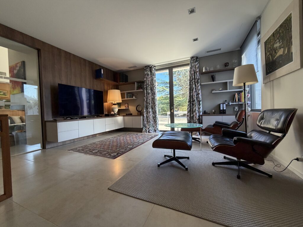 Agencement intérieur sur mesure d'un salon TV (projet particulier) par L'Arche du Bois. Un grand meuble TV moderne avec des éléments suspendus blancs et un habillage mural en bois fait face à des fauteuils en cuir noir. Sur la gauche, une porte vitrée coulissante sépare l'espace.