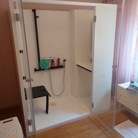Intérieur de la cabine de douche Danaé by L'Arche du Bois, portes ouvertes. L'unité est divisée : siège PMR et barres de maintien à gauche, et espace vasque/lavabo intégré à droite.