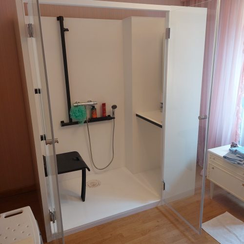 Intérieur de la cabine de douche Danaé by L'Arche du Bois, portes ouvertes. L'unité est divisée : siège PMR et barres de maintien à gauche, et espace vasque/lavabo intégré à droite.