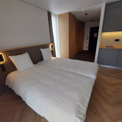 Chambre d'hôtel avec un lit double à tête de lit en tasseaux de bois, une kitchenette moderne grise et un sol en parquet en chevrons.
