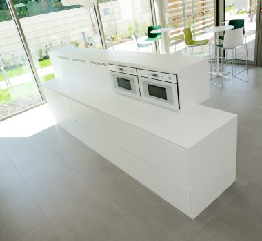 Cuisine sur mesure par LDC Agencement – L’Arche du Bois : îlot central blanc avec four et micro-ondes intégrés, tiroirs en bas, entouré de portes et fenêtres, avec deux petites tables et six tabourets.