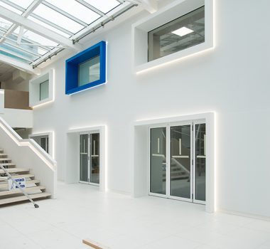 Vue d’ensemble d’un grand hall avec un escalier sur la gauche et un mur en face comportant plusieurs portes et fenêtres