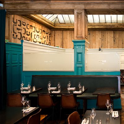 Intérieur de restaurant avec des tables et chaises de salle à manger, murs peints en bleu et plafond en bois apparent — un agencement chaleureux et contrasté signé Arche du Bois, alliant couleurs profondes et matériaux naturels.