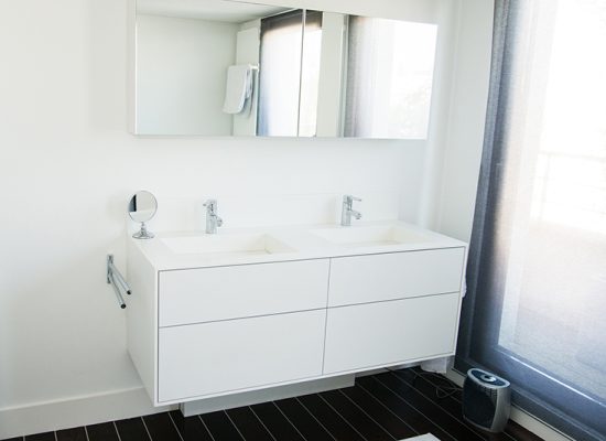 Salle de bain sur mesure par LDC Agencement – L’Arche du Bois : meuble vasque double avec deux robinets et miroir, porte vitrée à droite, design contemporain et agencement personnalisé.