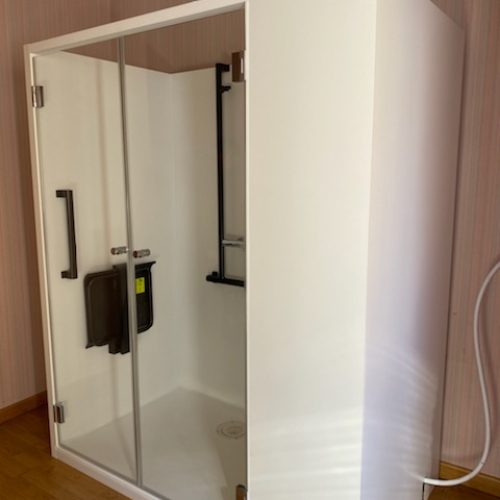 Cabine de douche Danaé by L'Arche du Bois vue de l'extérieur, portes fermées. À travers le verre vitré, le siège PMR rabattable et la barre de maintien verticale en I sont visibles.