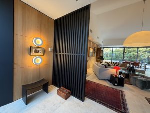 Agencement intérieur sur mesure d'une entrée moderne (projet particulier) par L’Arche du Bois à Eygalières. Un habillage en panneaux de hêtre avec deux appliques lumineuses fait face à un grand claustra en bois noir ajouré laissant entrevoir le salon.