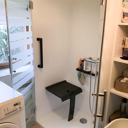 Installation de la douche accessible Danaé by L'Arche du Bois, portes ouvertes, avec intérieur blanc, siège PMR et barre d'appui en I, positionnée à côté d'une machine à laver.