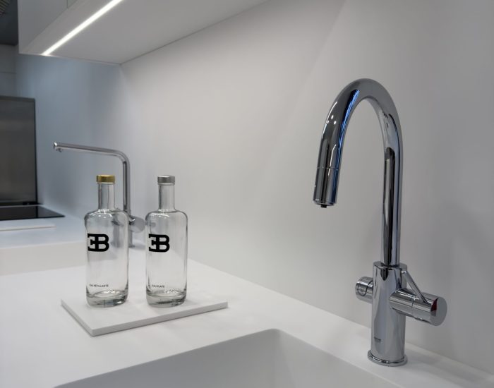 projet-cuisine-haut-de-gamme-bugatti-design-minimaliste Détail d'un évier blanc épuré avec robinetterie chromée et bouteilles siglées EB dans une cuisine professionnelle au design minimaliste pour le projet cuisine Bugatti.