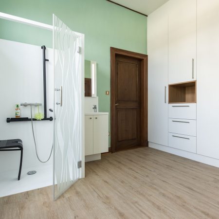 Cabine de douche Danaé by L'Arche du Bois, portes ouvertes, installée dans une pièce d'habitation. La cabine tout-en-un intègre siège PMR, double barre d'appui, vasque et miroir, à côté d'une porte en bois et d'une bibliothèque.