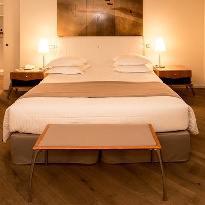 Lit double avec deux oreillers, un bout de lit placé en face, un cadre mural au-dessus de la tête de lit et deux lampes de chevet positionnées de chaque côté — un agencement harmonieux et confortable signé Arche du Bois.