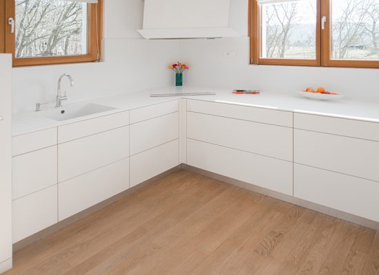 Vue d’angle d’une cuisine moderne montrant des armoires murales des deux côtés, avec un évier positionné à gauche sous une fenêtre. Deux fenêtres situées au-dessus des armoires assurent une belle luminosité naturelle.