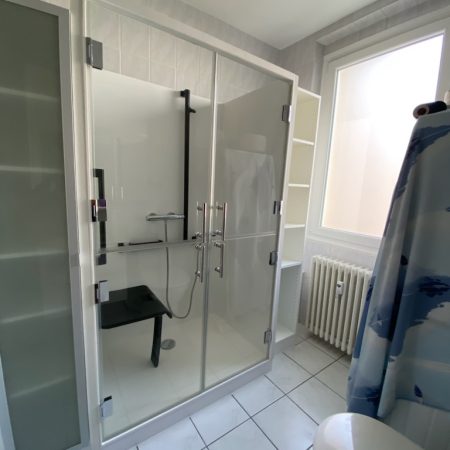 Vue extérieure de la cabine de douche Danaé by L'Arche du Bois avec portes fermées en verre vitré, laissant apercevoir le siège PMR et la double barre d'appui (en I et en T).