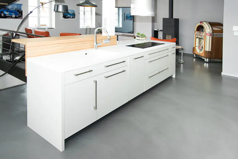 Cuisine sur mesure par LDC Agencement – L’Arche du Bois : îlot central blanc avec tiroirs et rangements intégrés, équipé d’un lavabo avec robinet et d’une plaque de cuisson sur le plan de travail, chaises et abat-jour en arrière-plan avec deux fenêtres.